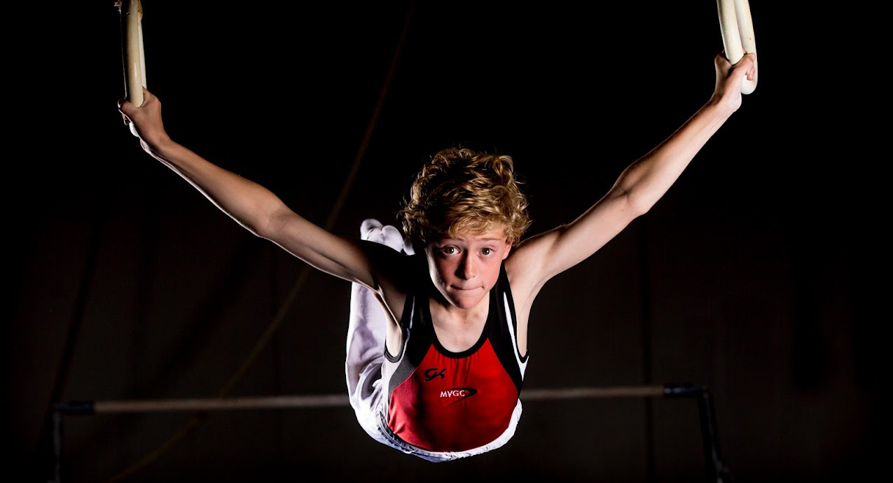 boy gymnast swinging from rings black background, wearing MYGC red uniform