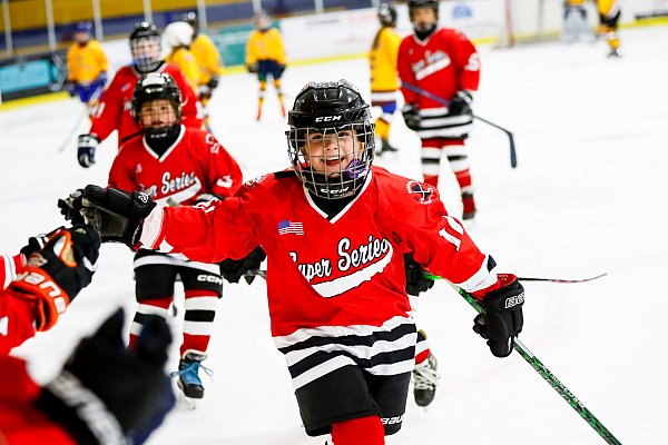 young boy hockey player in red jersey gives high five to players on bench while smiling at the camera during hockey game